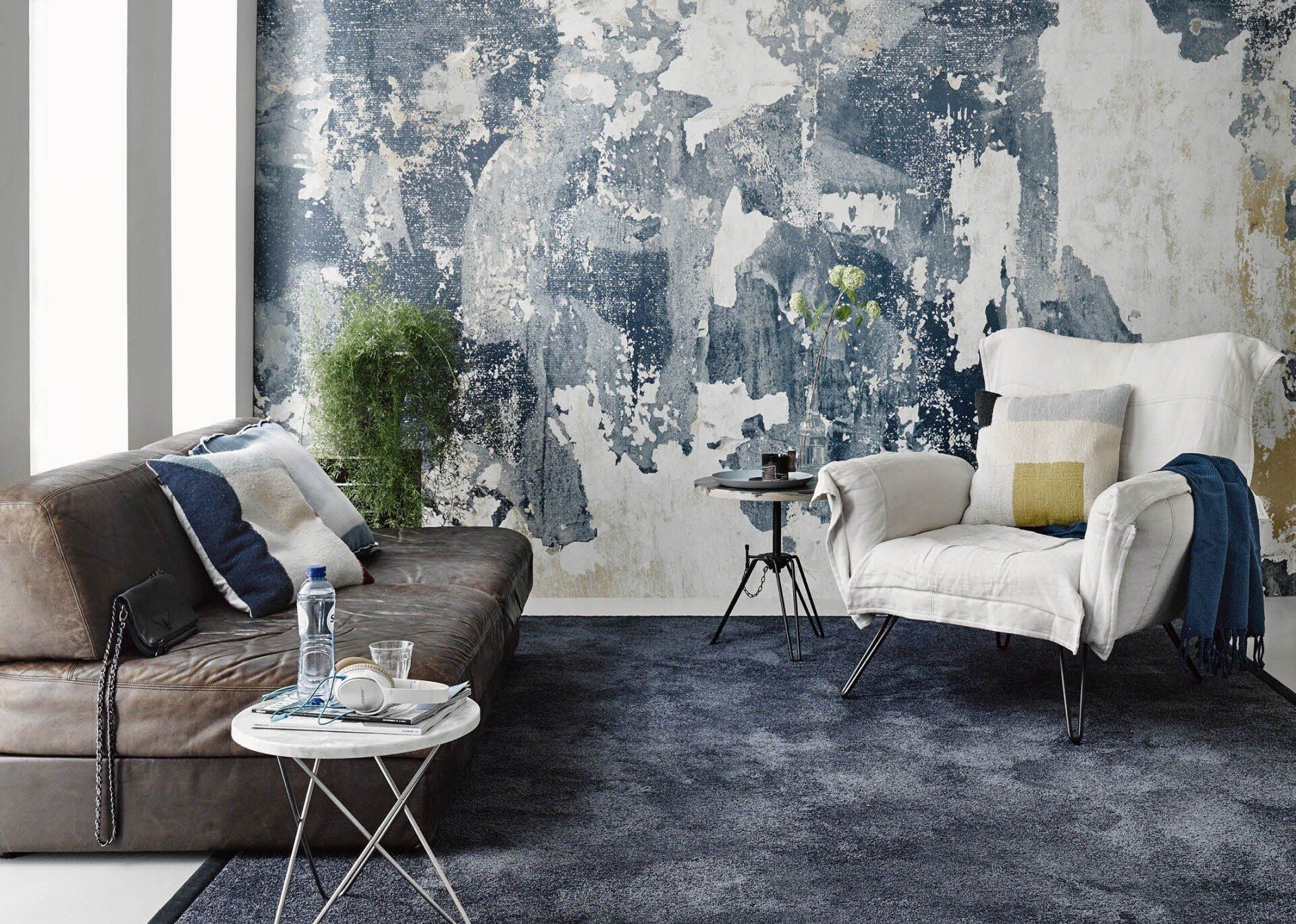 Woonkamer met verweerde blauw-witte muur, bruine bank, witte fauteuil en donkerblauw vloerkleed.