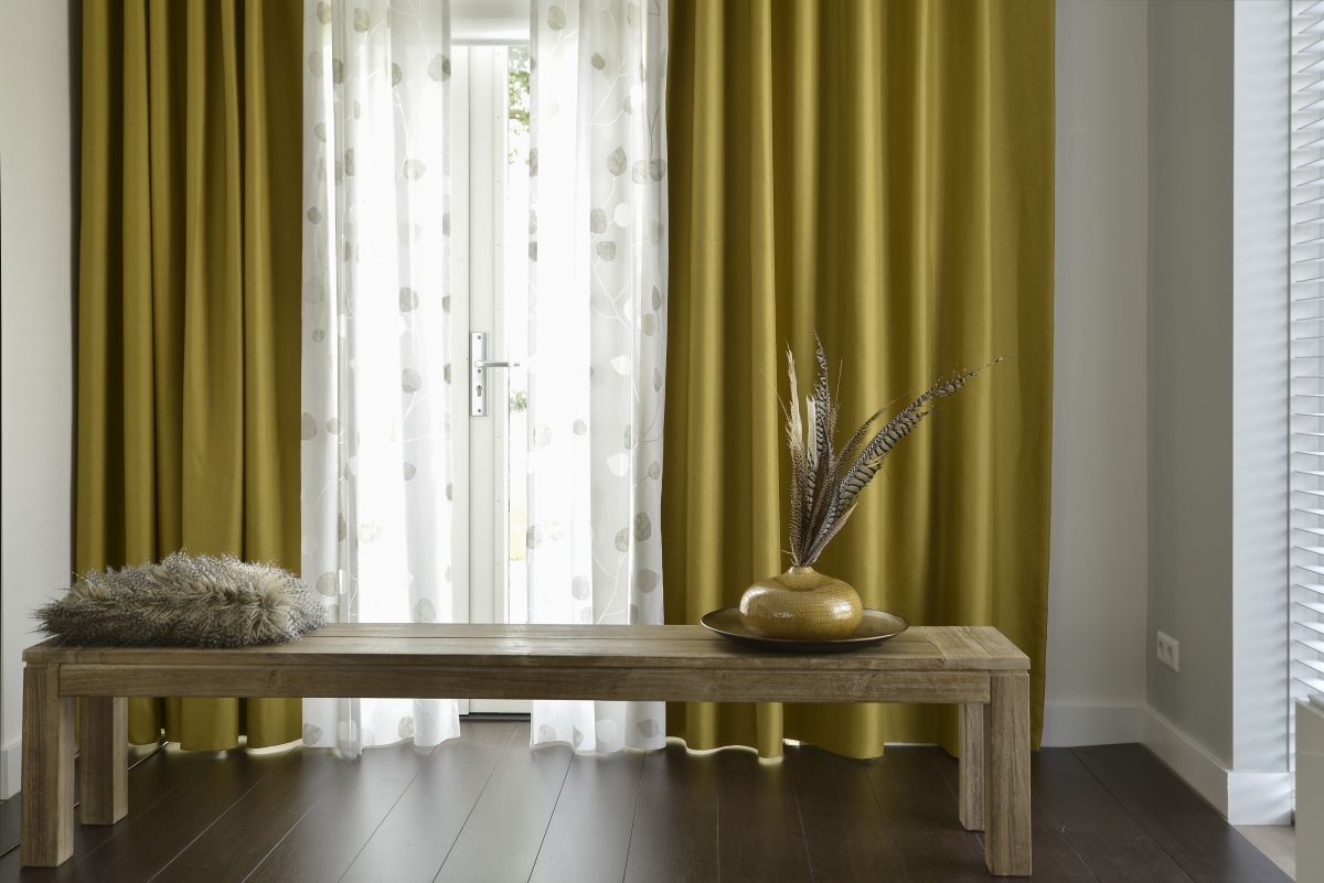 Mosterdkleurige gordijnen omlijsten een raam met transparante witte panelen; houten bank met decoratie.