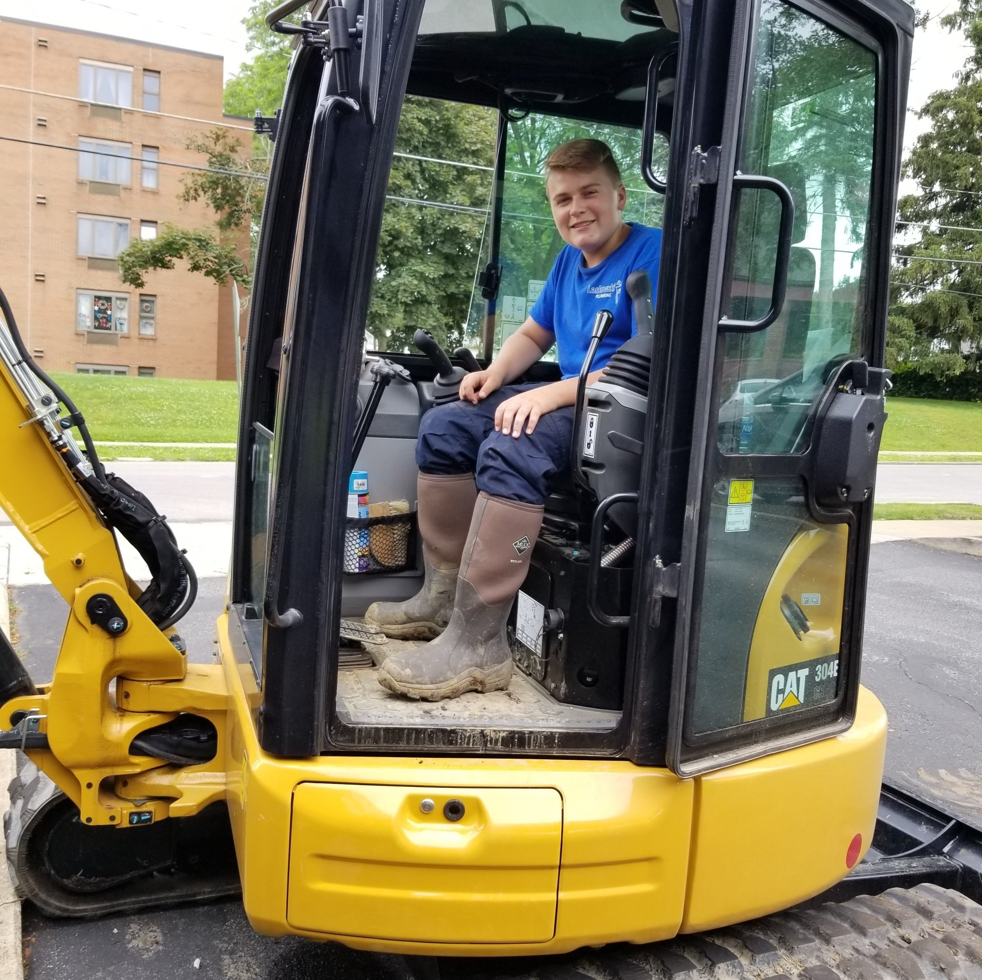 Young man in blue shirt and boots in the cab of a yellow CAT excavator.