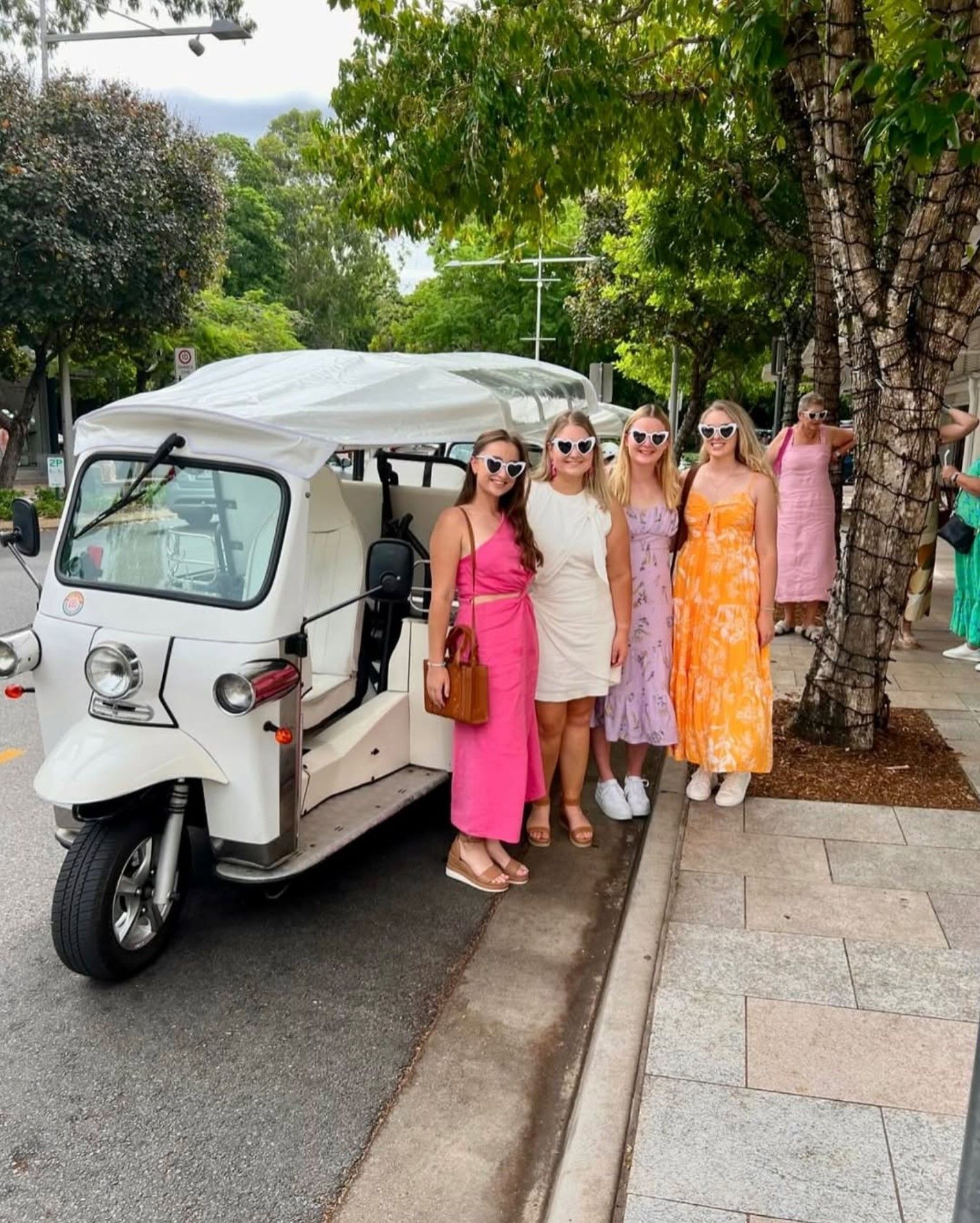 Five women in dresses pose near a white tuk-tuk on a sidewalk lined with trees.