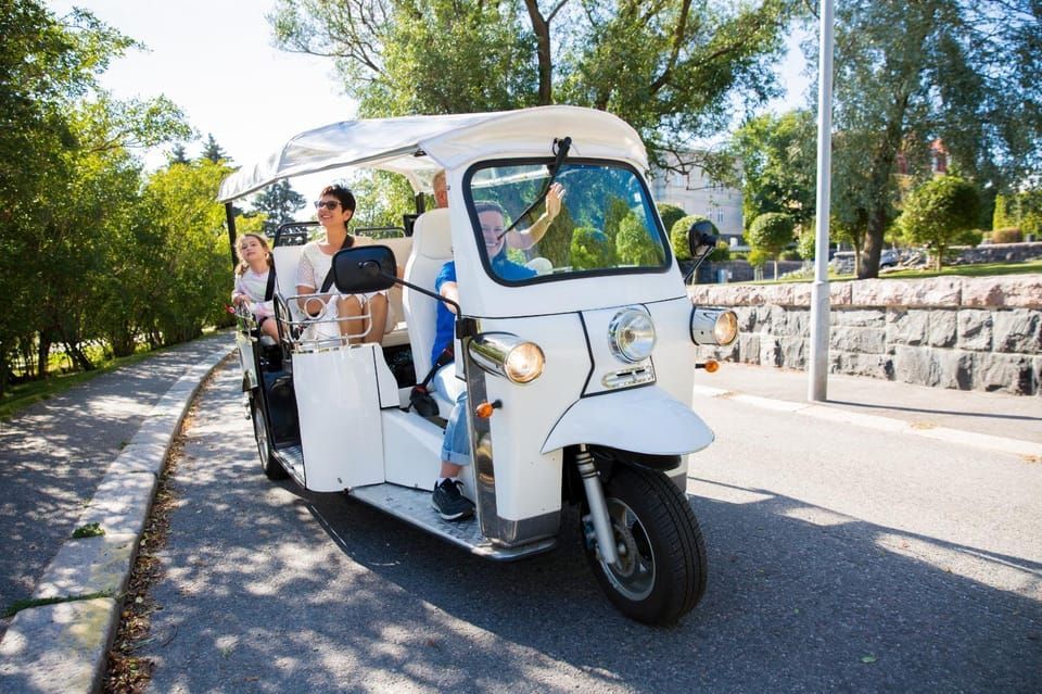 White tuk-tuk with passengers driving on a paved road next to a green space.