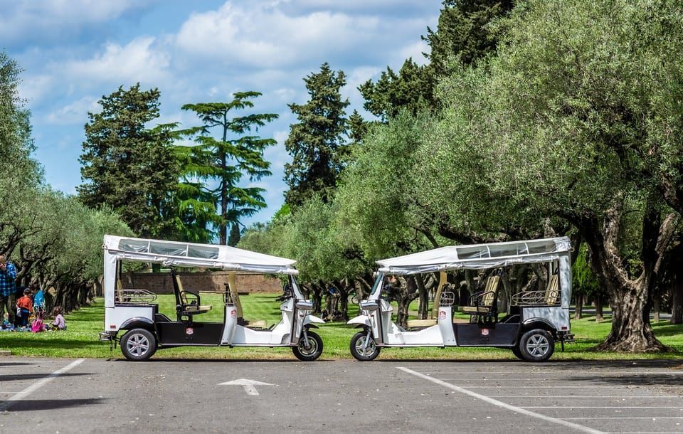Two white electric Tuk Tuk tour vehicles parked on asphalt, green trees in background.