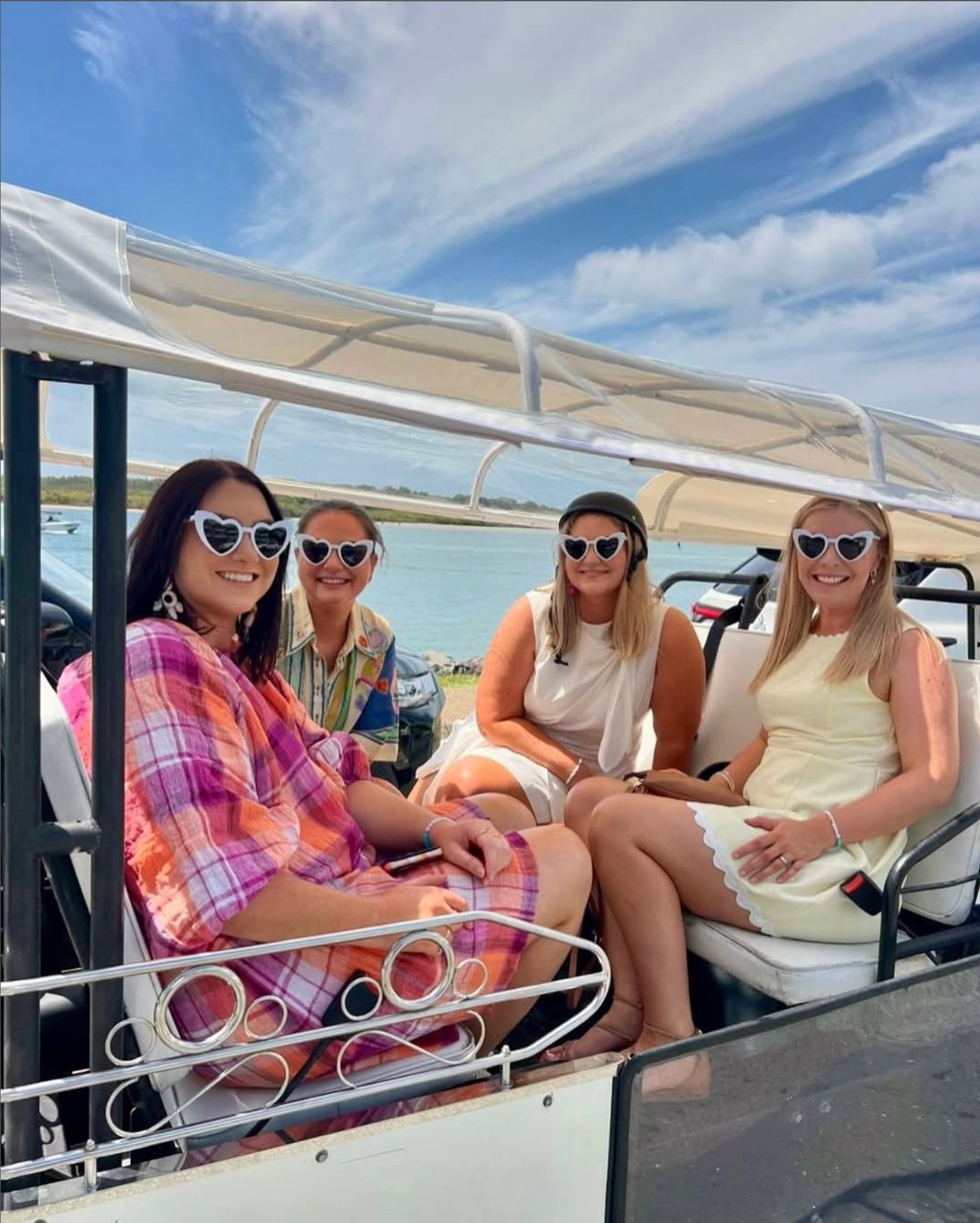 Four people in sunglasses ride a white golf cart on a sunny day near water.