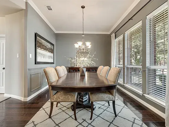 Bright dining room with a long wooden table, cream chairs, chandelier, and large windows with blinds