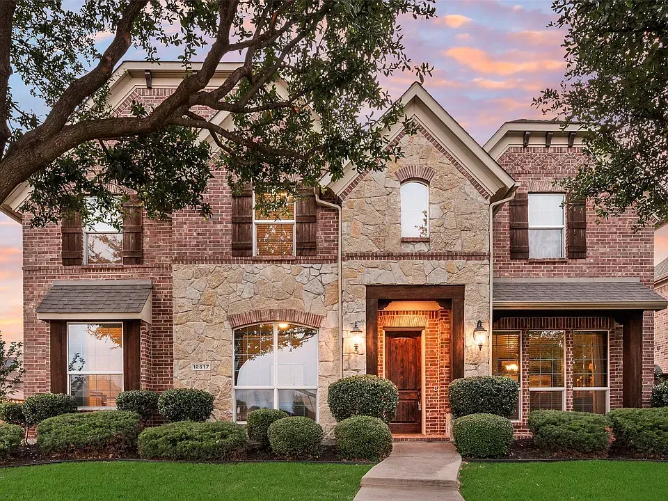 Large brick suburban house with stone facade, lit front door, and manicured shrubs at sunset