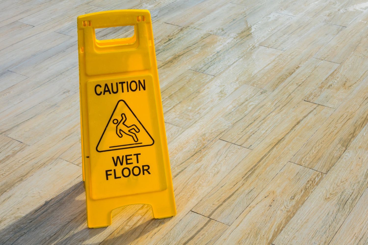 Yellow caution sign on a wooden floor, warning of a wet floor with a graphic of a person slipping.