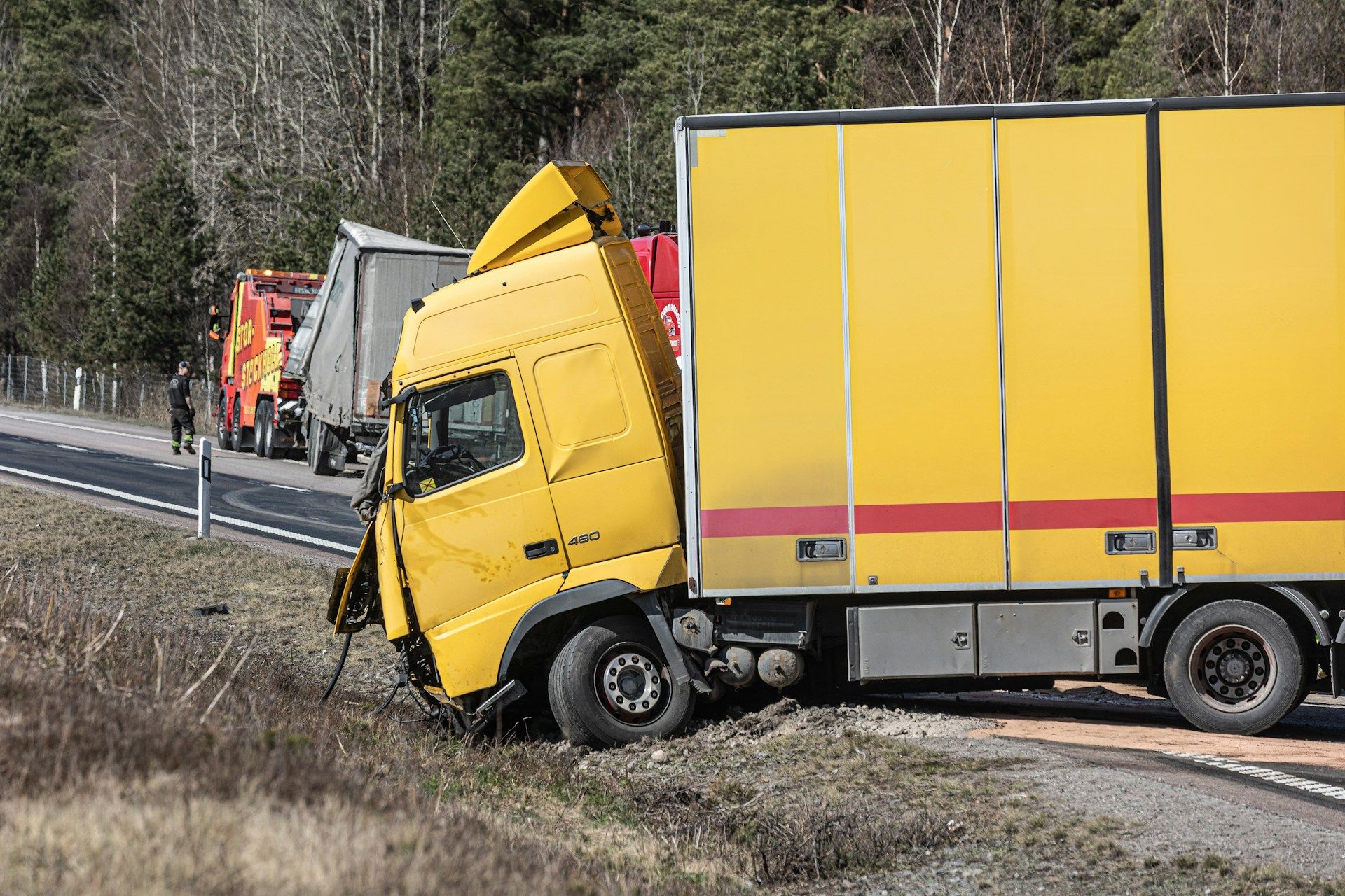 Yellow semi-truck crashed off road, partially blocking a highway. Rescue vehicle and people present.