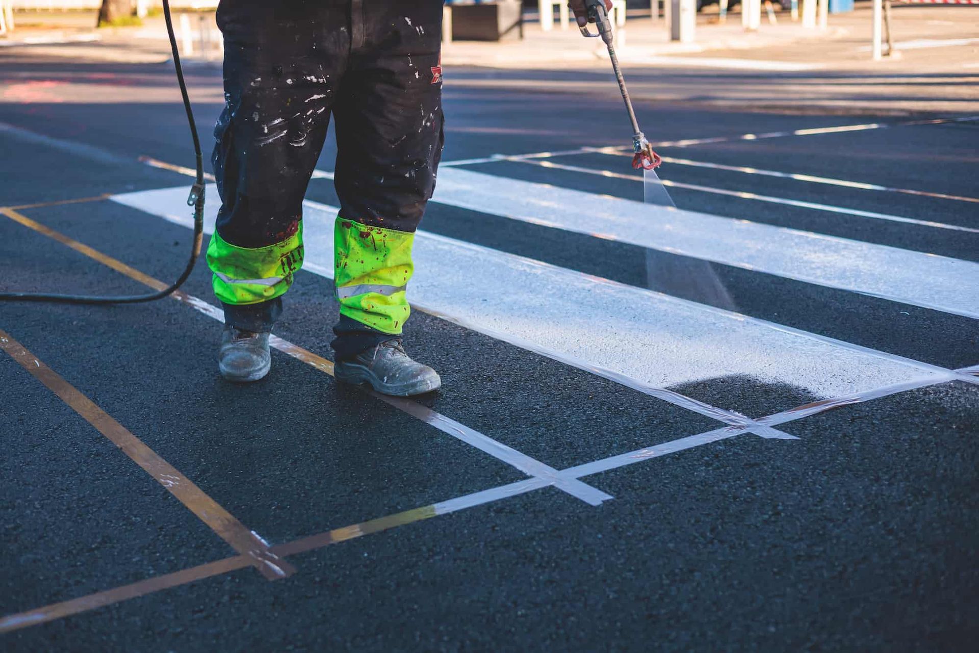 Person painting a crosswalk, using a spray gun. Legs in protective gear, white paint on the dark asphalt.