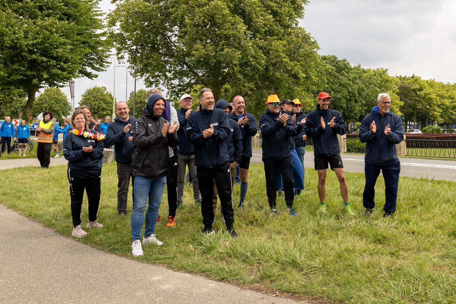 Een groep mensen in regenjassen juicht buiten langs een pad en een grasveld onder een bewolkte hemel.