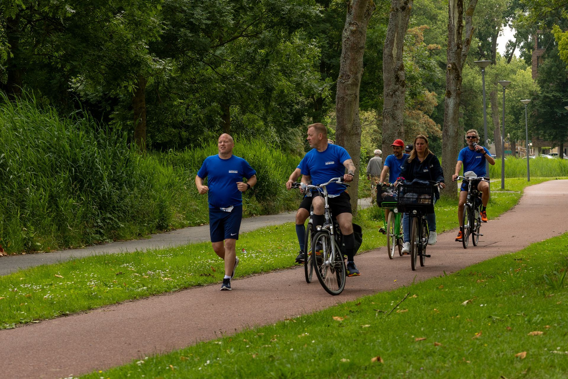 Fietsers en een hardloper op een met bomen omzoomd parkpad op een regenachtige dag.