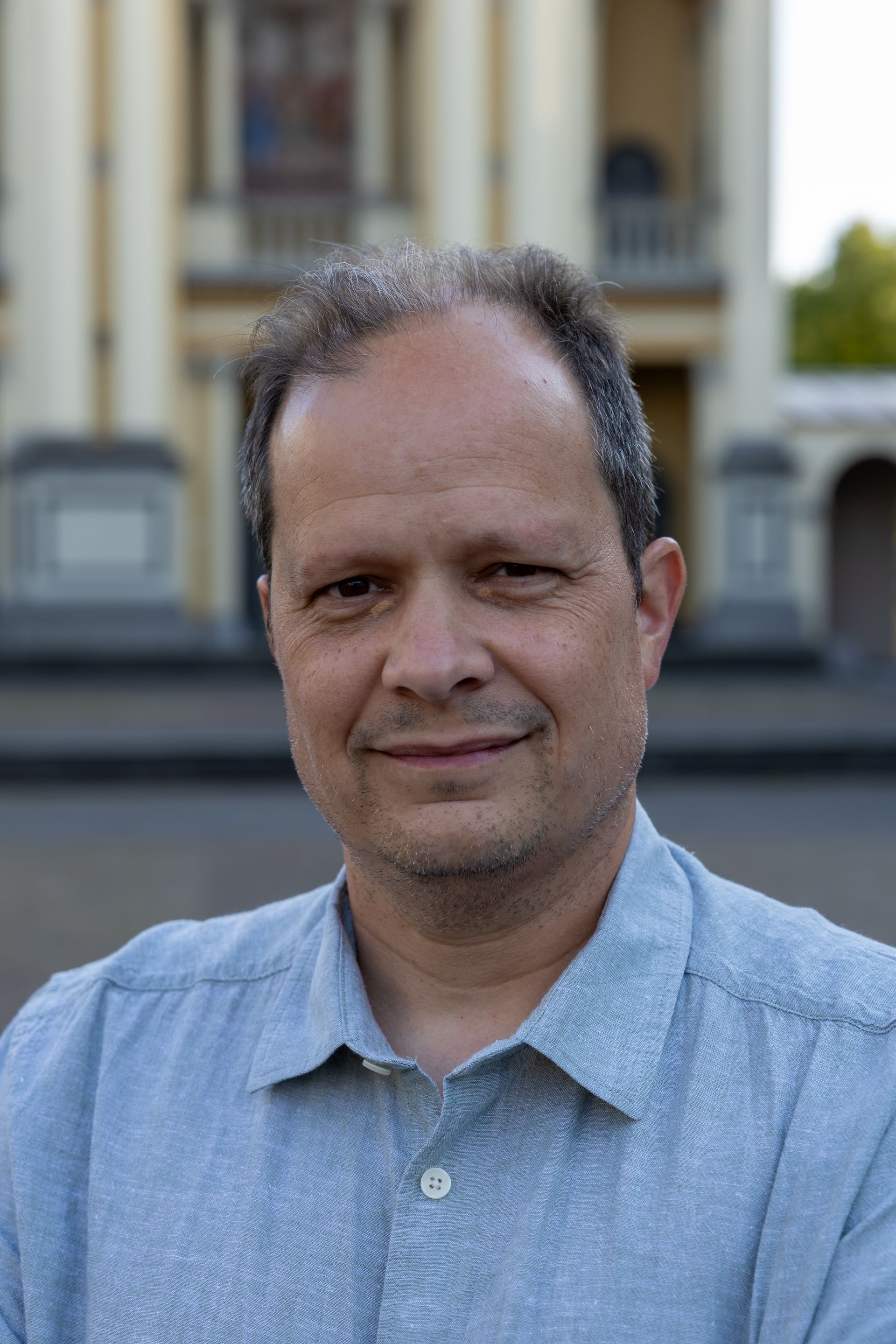 Portret van een man in een lichtblauw shirt, staand in de buitenlucht met een gebouw dat wazig op de achtergrond te zien is.