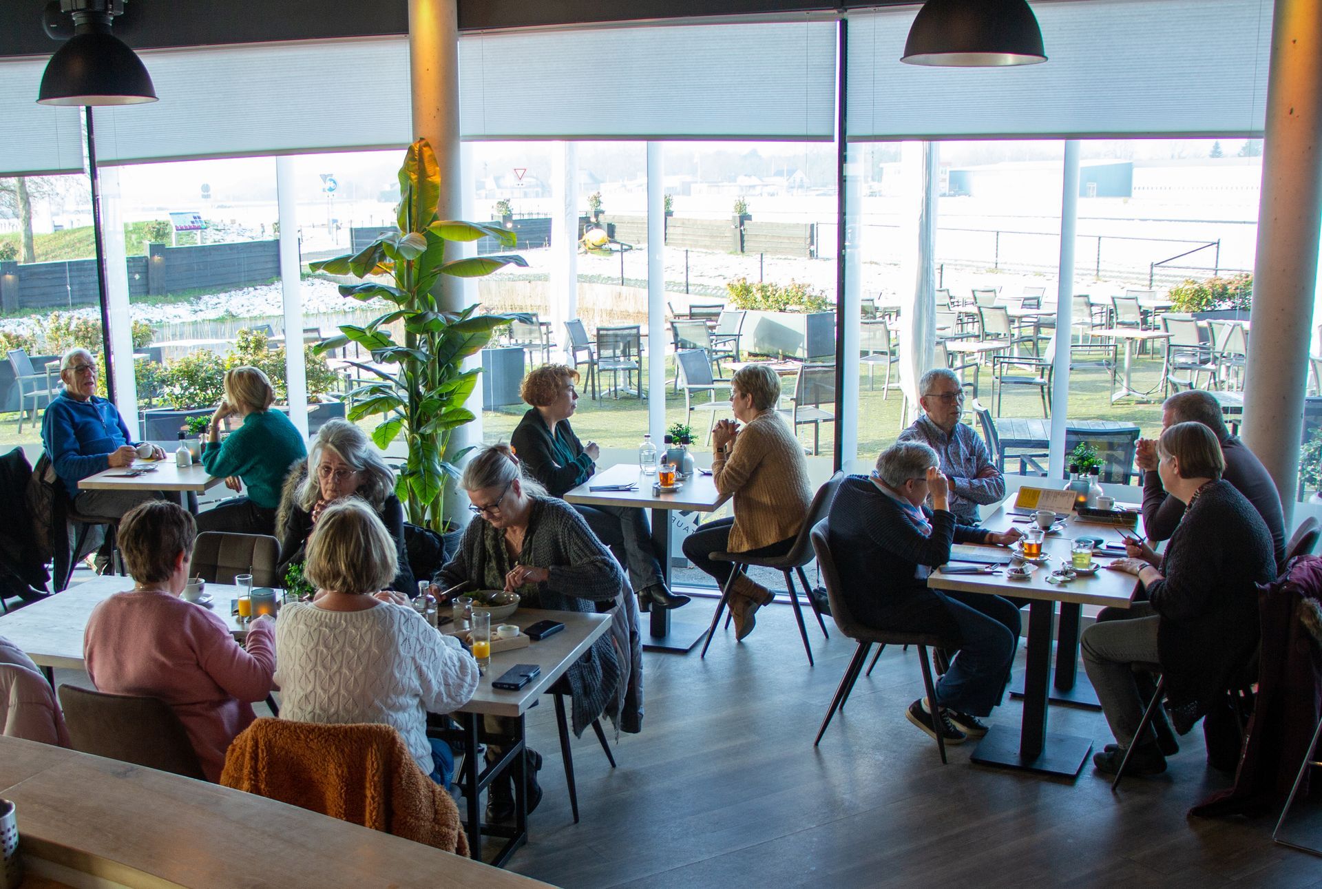 Mensen dineren aan tafels in een licht café met grote ramen en hangende lampen.