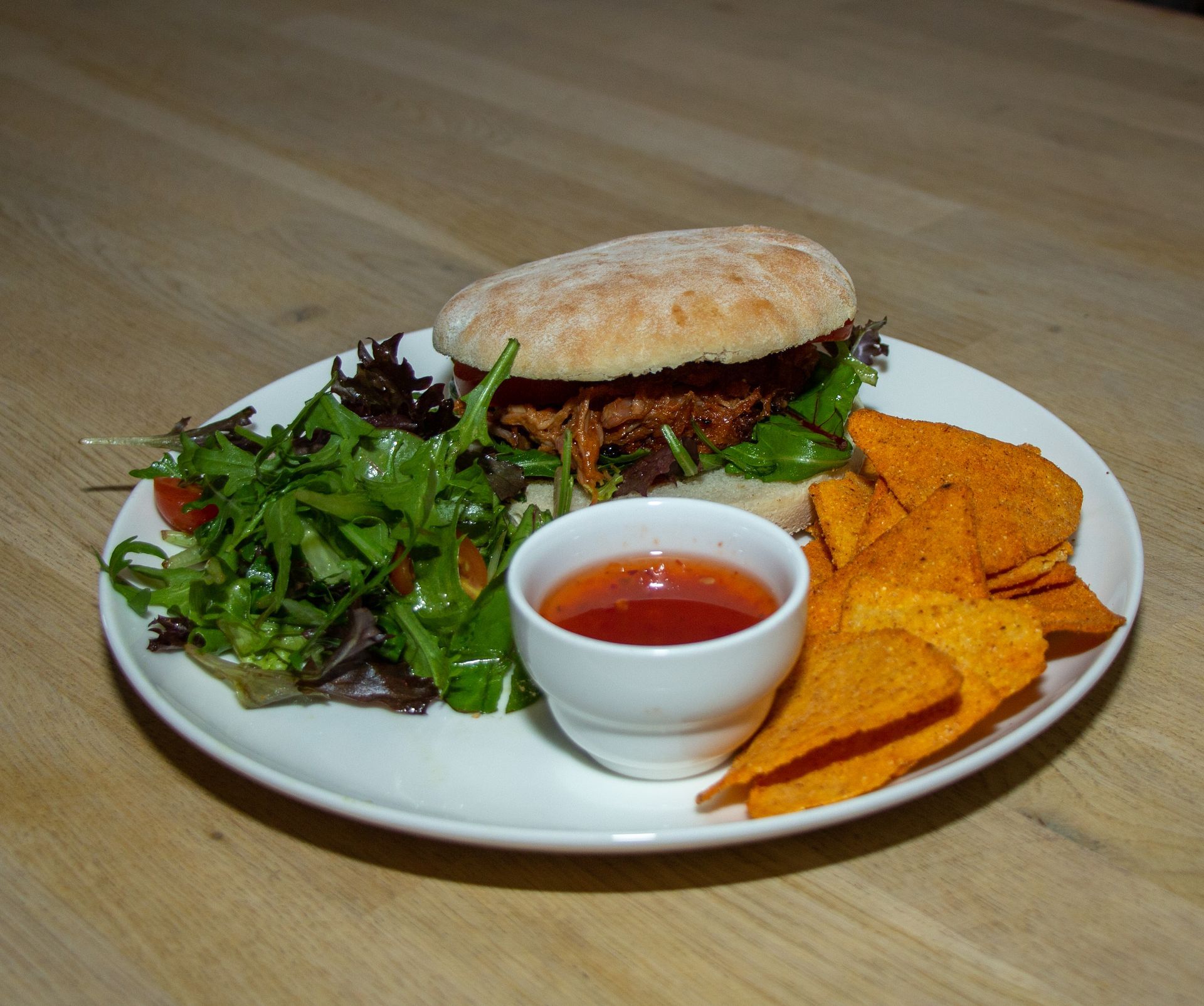 Een sandwich met groene salade, tortillachips en een klein bakje rode dipsaus op een wit bord.