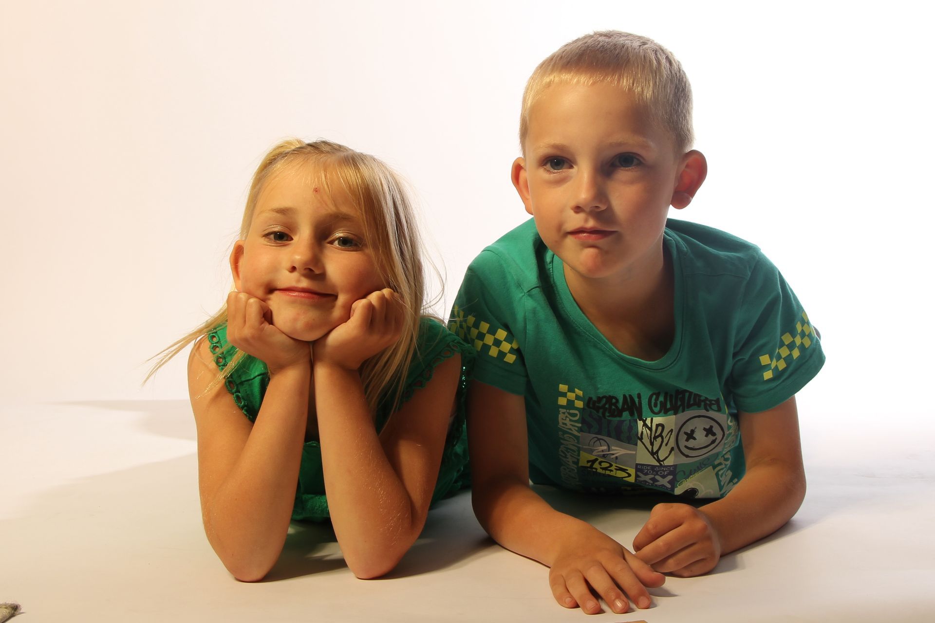 Twee kinderen liggen op een witte ondergrond, de ene glimlacht met zijn kin op zijn handen, de andere kijkt recht vooruit.