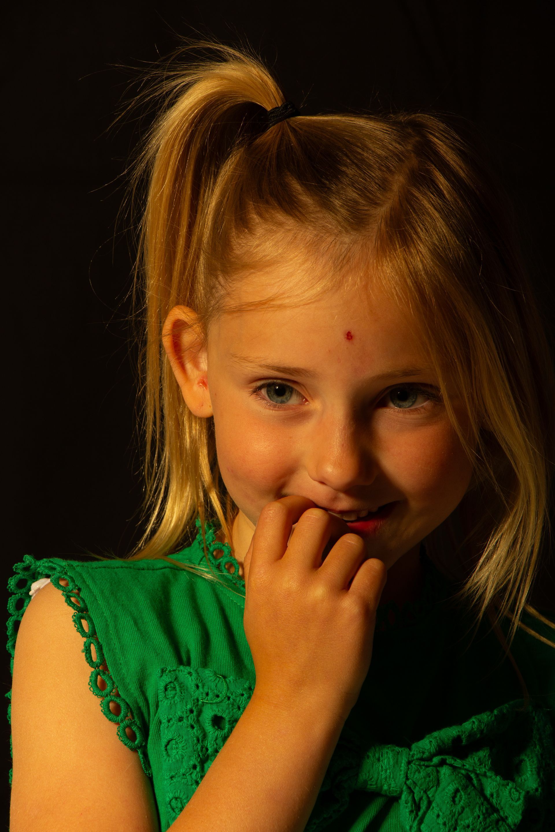 Lachend kind in een groene jurk met hand bij de mond tegen een donkere achtergrond.
