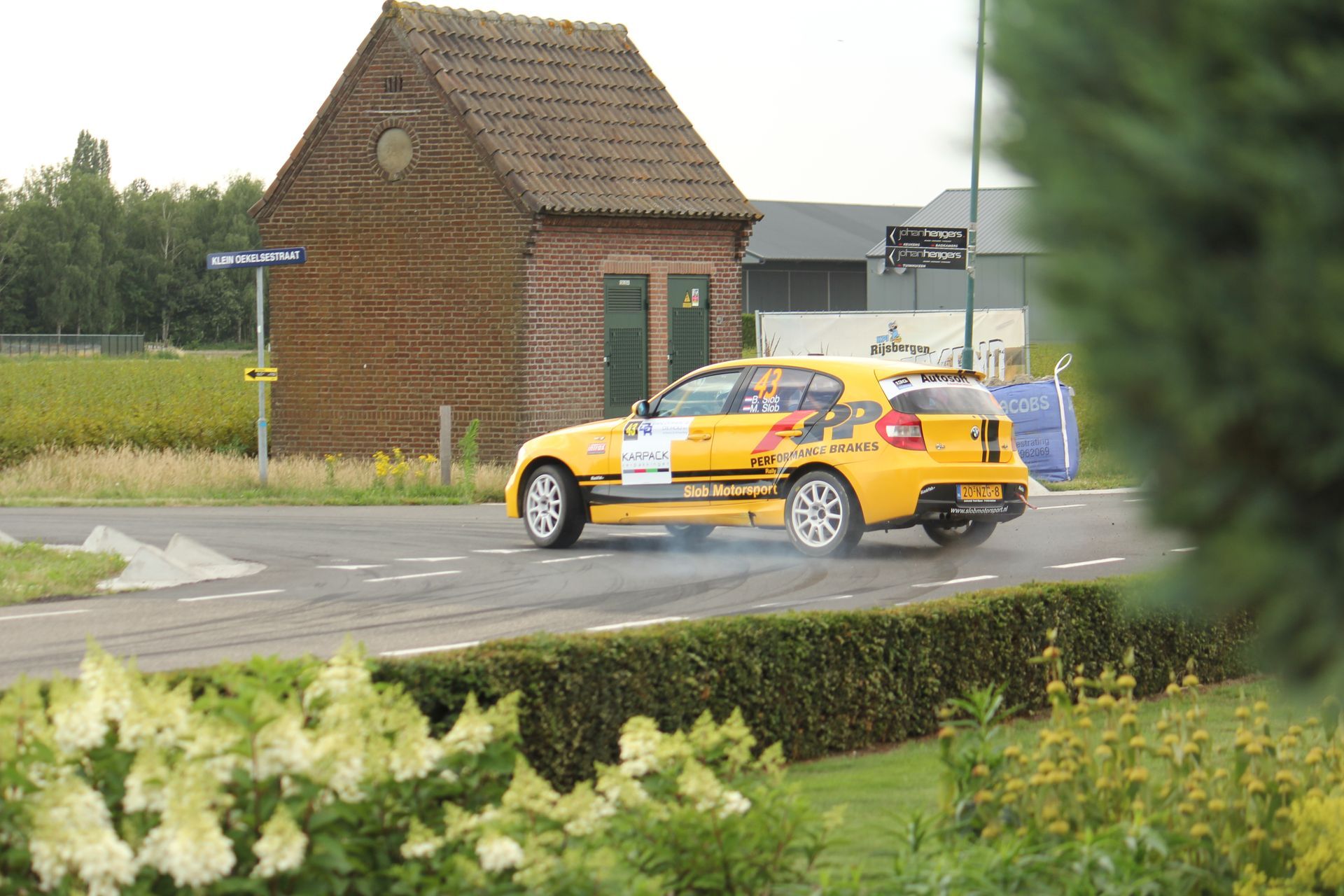 Gele raceauto op een natte weg vlakbij een bakstenen gebouw, gedeeltelijk omgeven door groen.