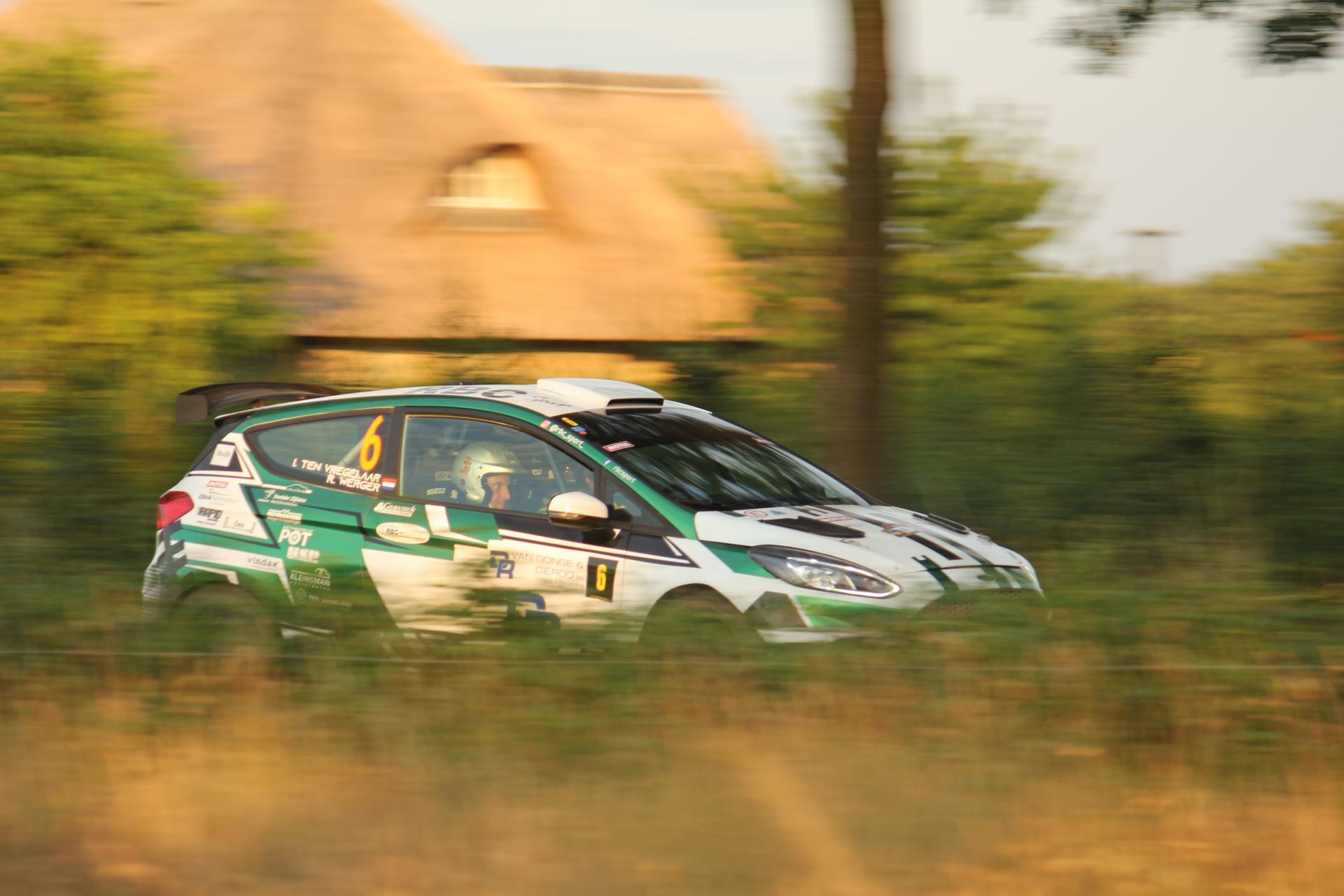 Groene rallyauto die door hoog gras raast, met een wazige achtergrond en stofspoor.