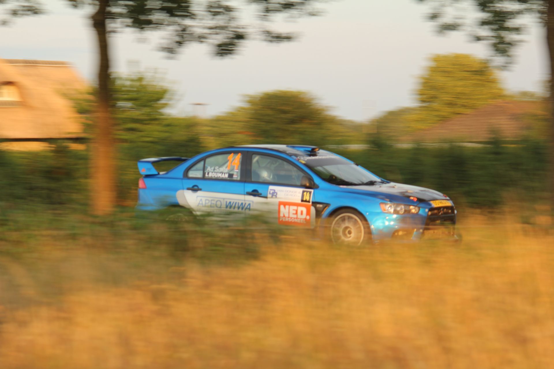 Een blauwe rallyauto raast door een grasveld bij zonsondergang.