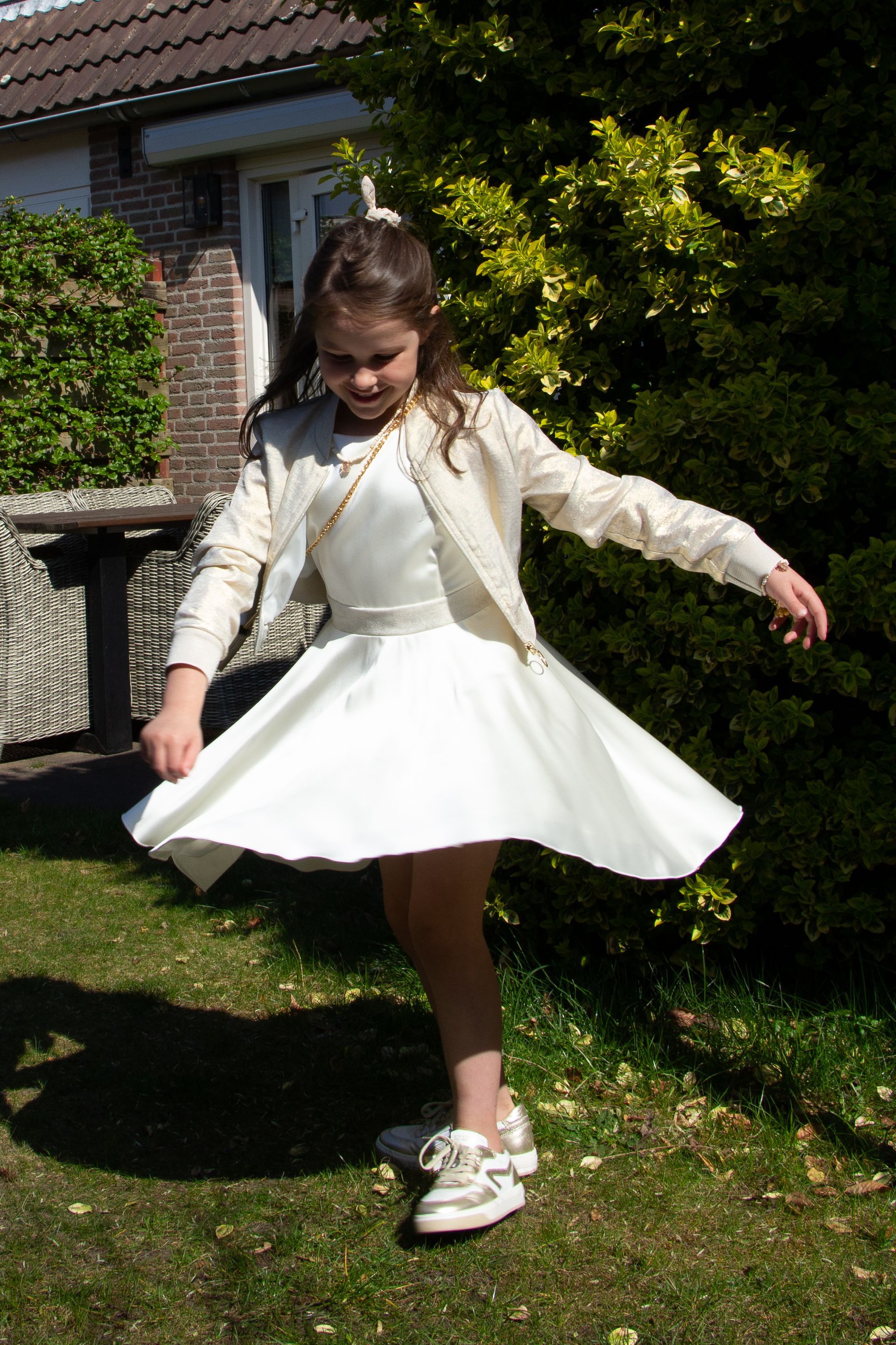 Meisje in een witte jurk en jasje draait rond op een gazon voor een huis.