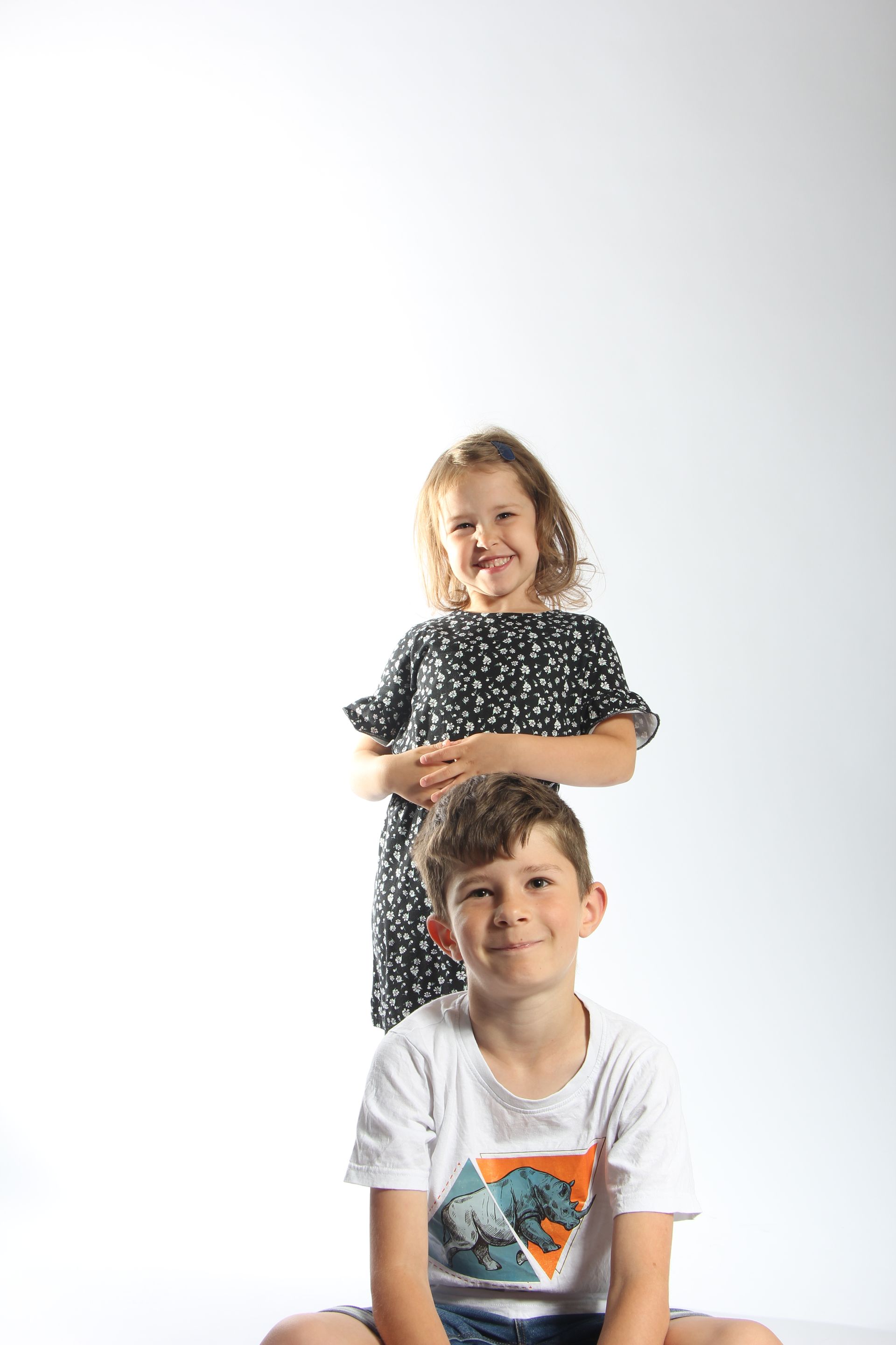 Twee kinderen poseerden voor een witte achtergrond, de ene achter de andere, beiden lachend naar de camera.