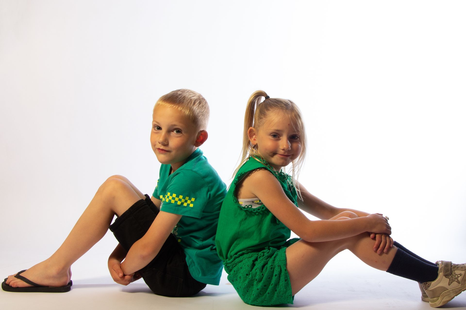 Twee kinderen in groene kleding zitten rug aan rug op een witte studiovloer.