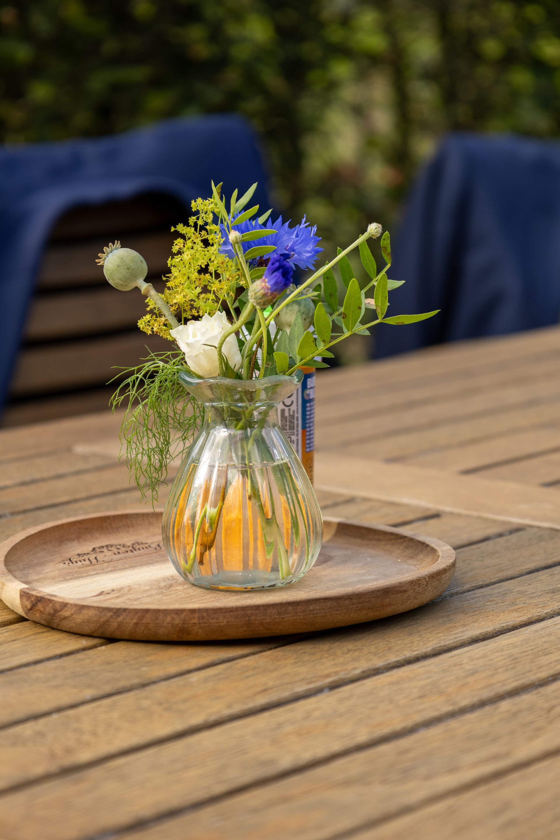 Klein boeketje wilde bloemen in een glazen vaas op een houten tafel met blauwe stoelen op de achtergrond.