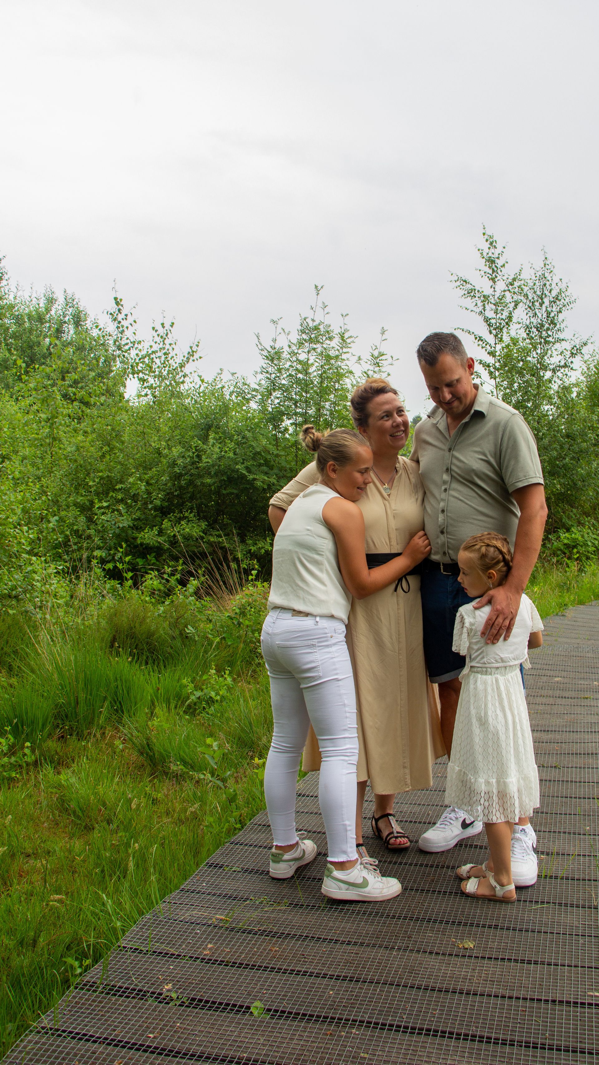 Een gezin staat samen op een houten pad naast weelderige groene struiken onder een bewolkte hemel.