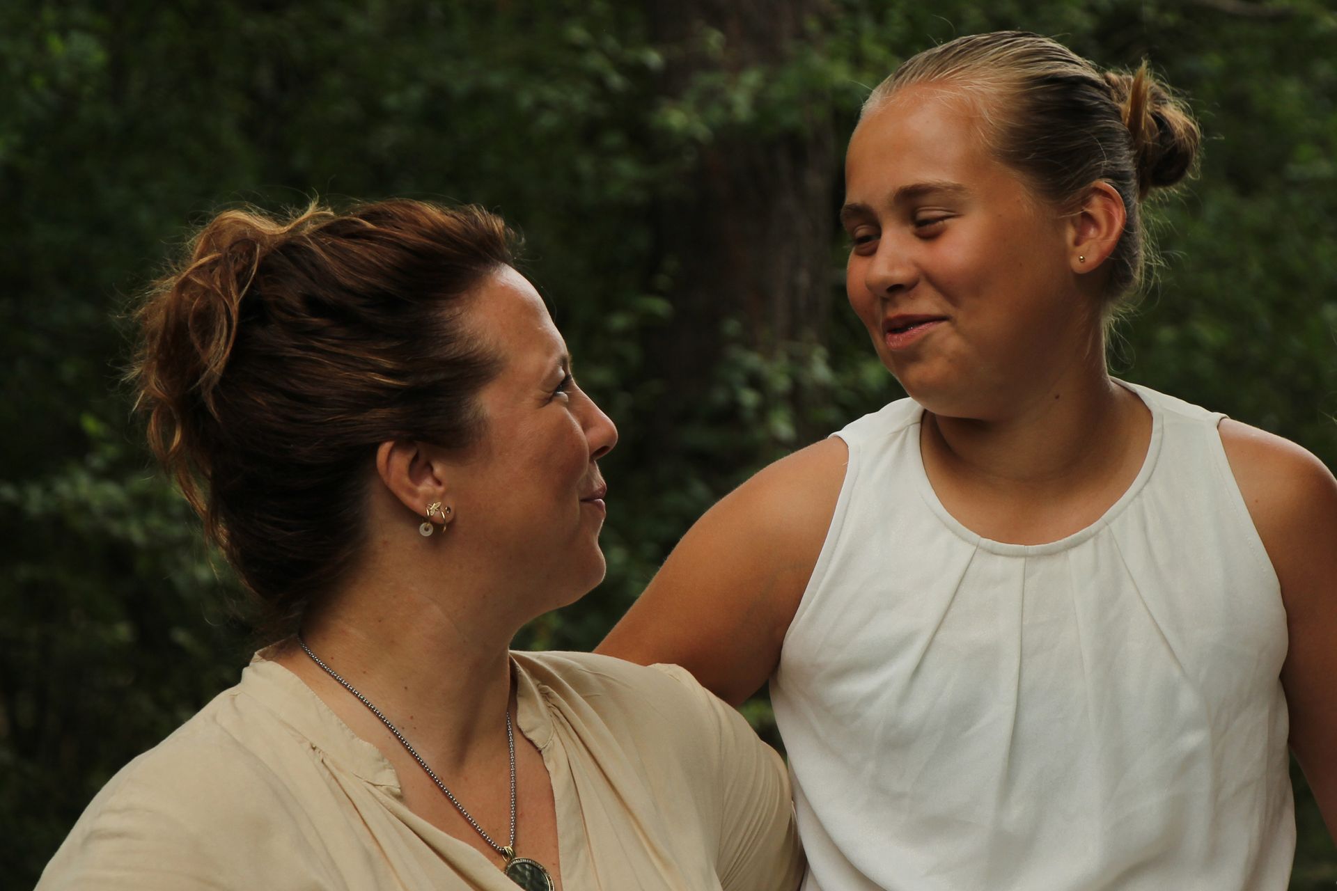Twee vrouwen glimlachen naar elkaar buiten, de ene in een witte top en de andere in een beige top.