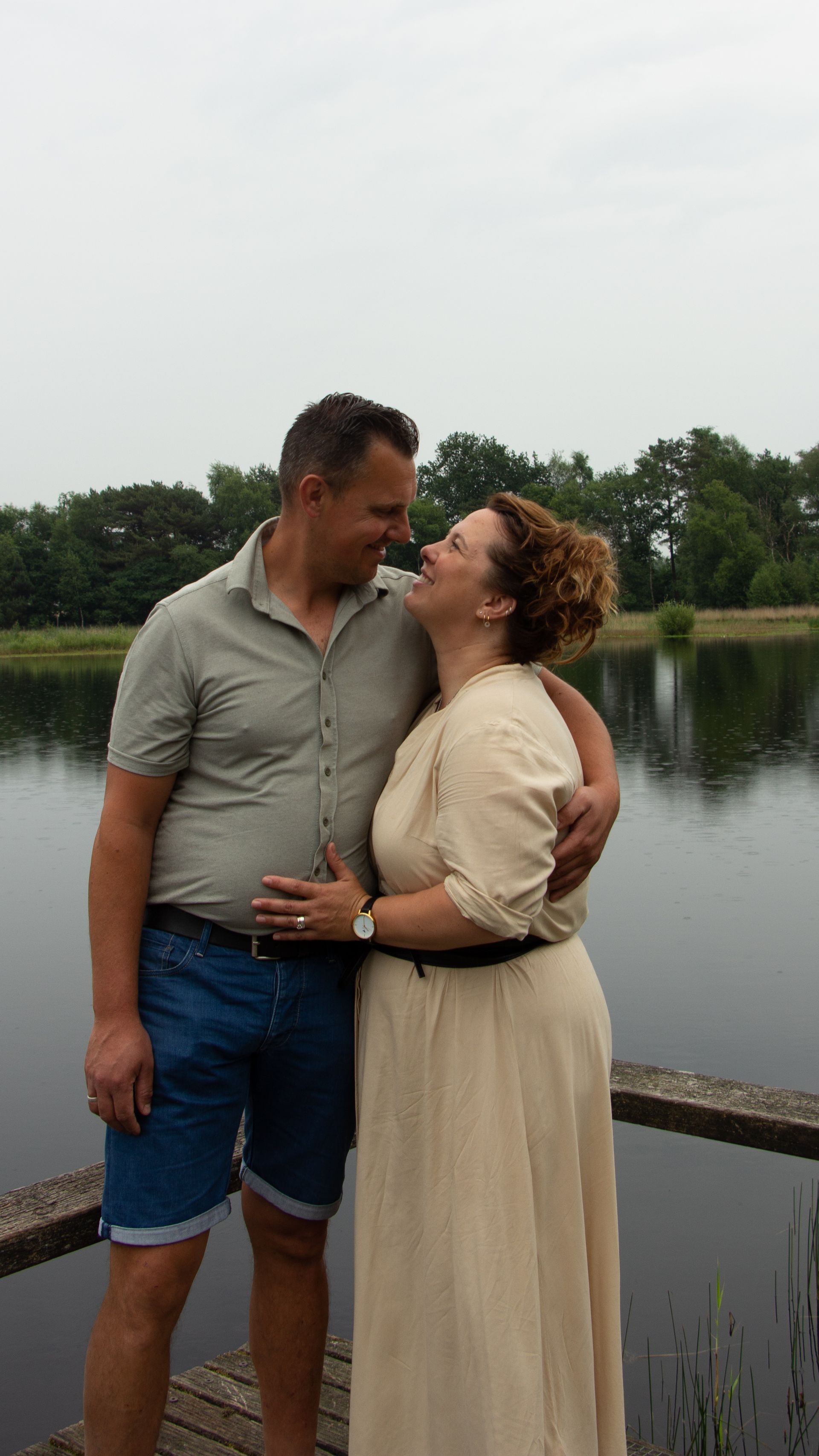 Een stel omhelst elkaar bij een reling aan het meer en lacht elkaar toe in casual zomerkleding.