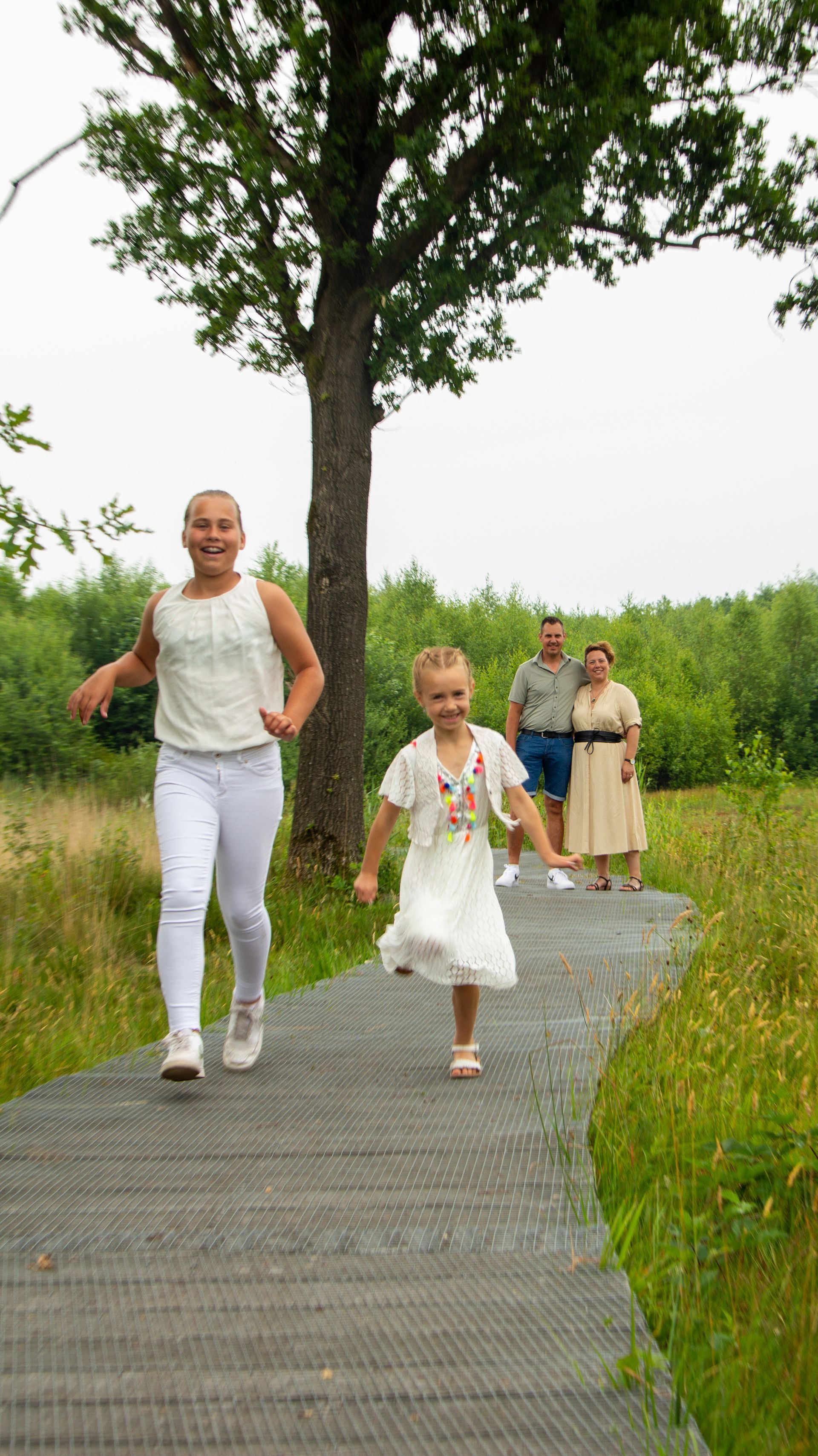 Mensen lopen over een houten pad door een groen veld, aangevoerd door een lachende vrouw in het wit.