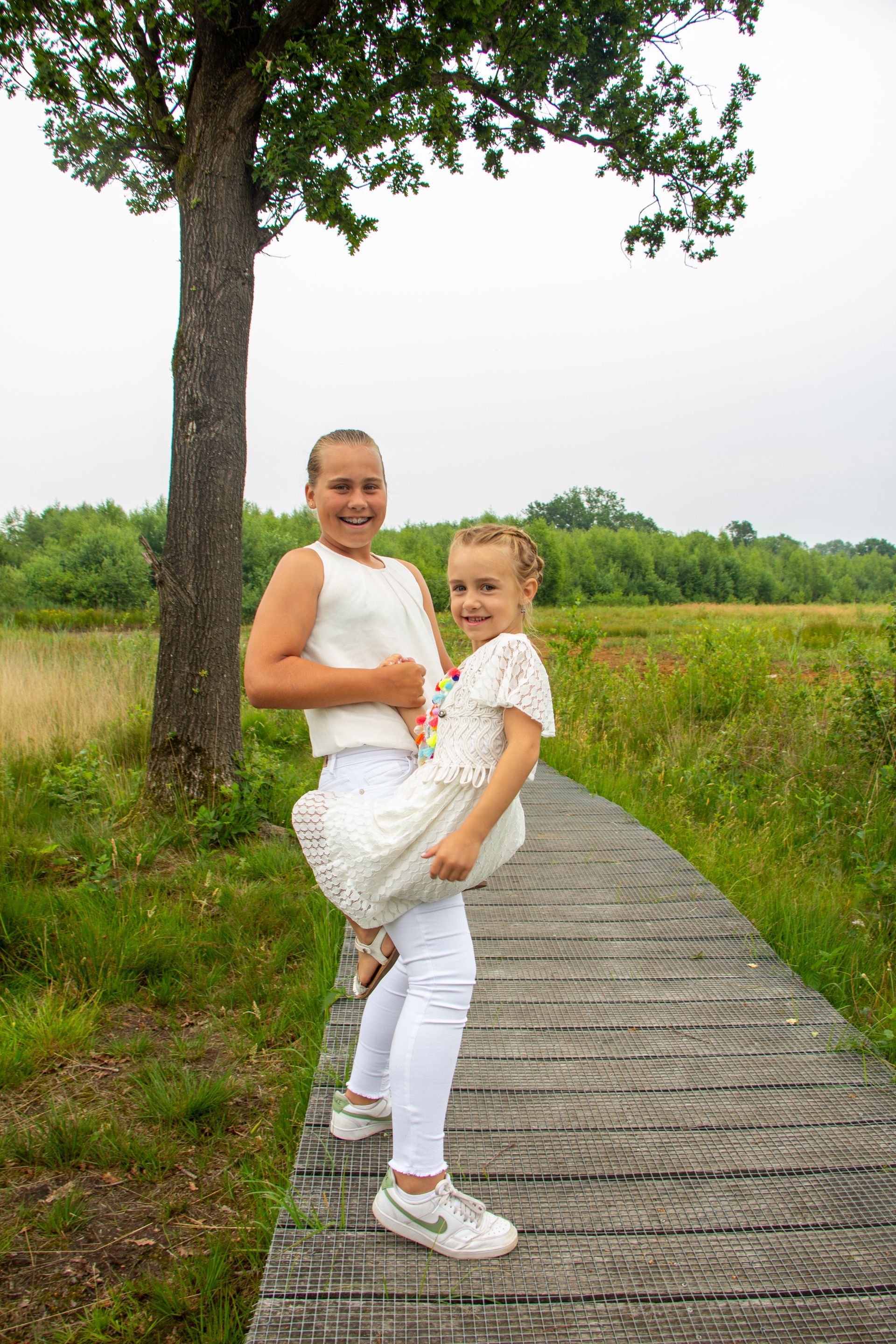 Twee mensen in witte kleding staan op een houten pad bij een boom in een grasveld en glimlachen naar de camera.