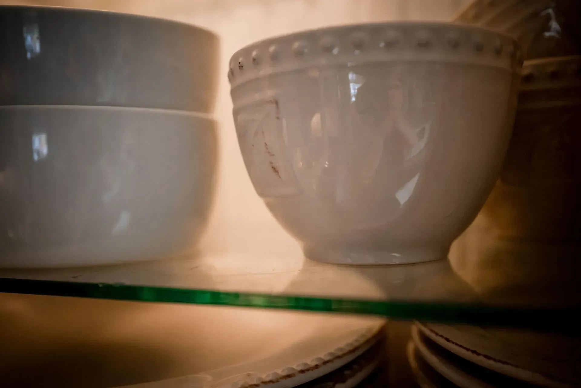Two white bowls are sitting on a glass shelf next to a stack of plates