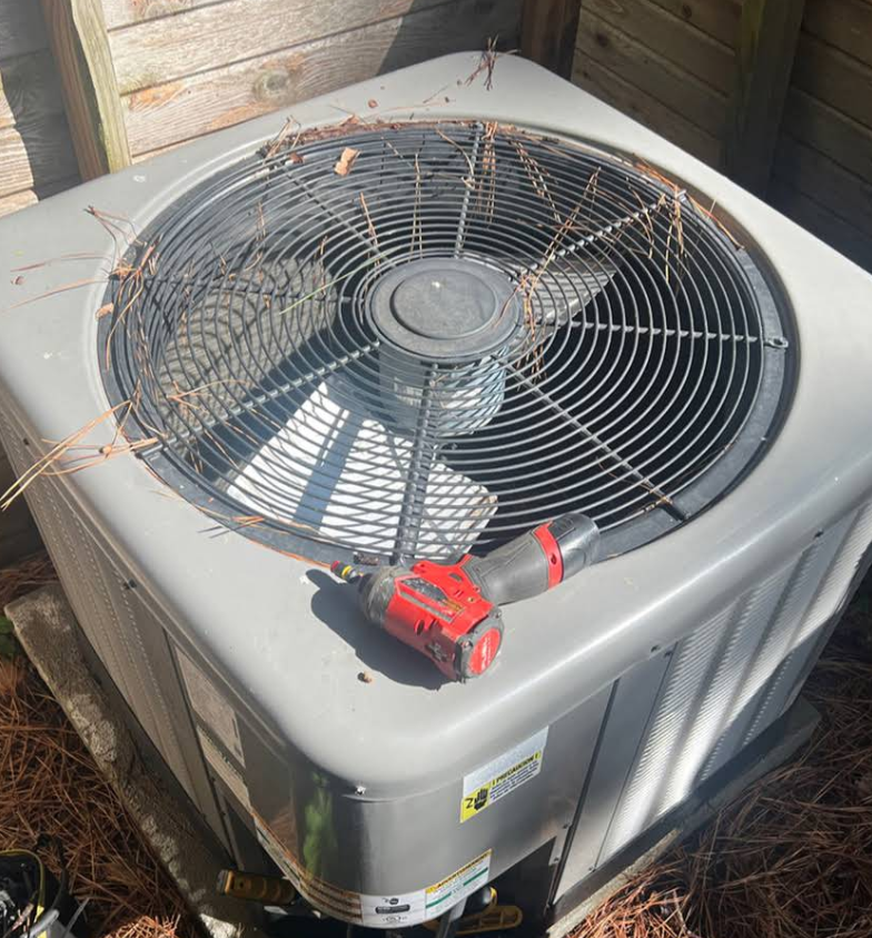 Outdoor air conditioner unit with a red power tool resting on top, surrounded by debris and leaves