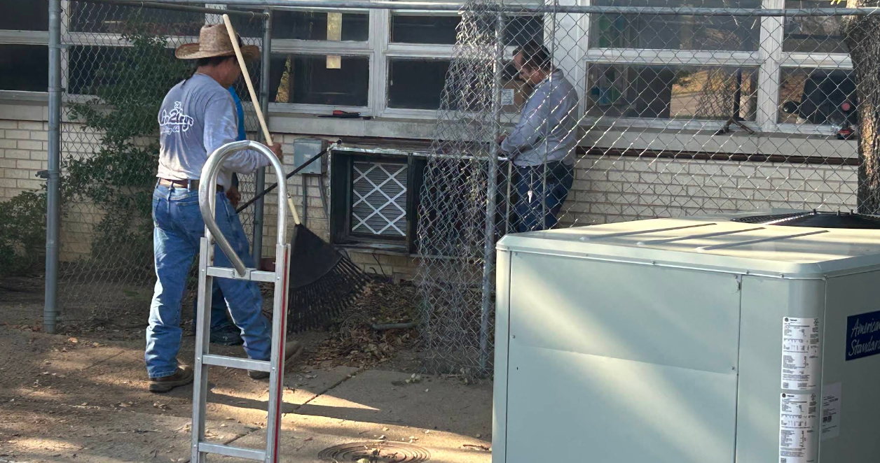 Two workers in blue shirts near a chain-link fence, with a step ladder and utility box in the foreground.