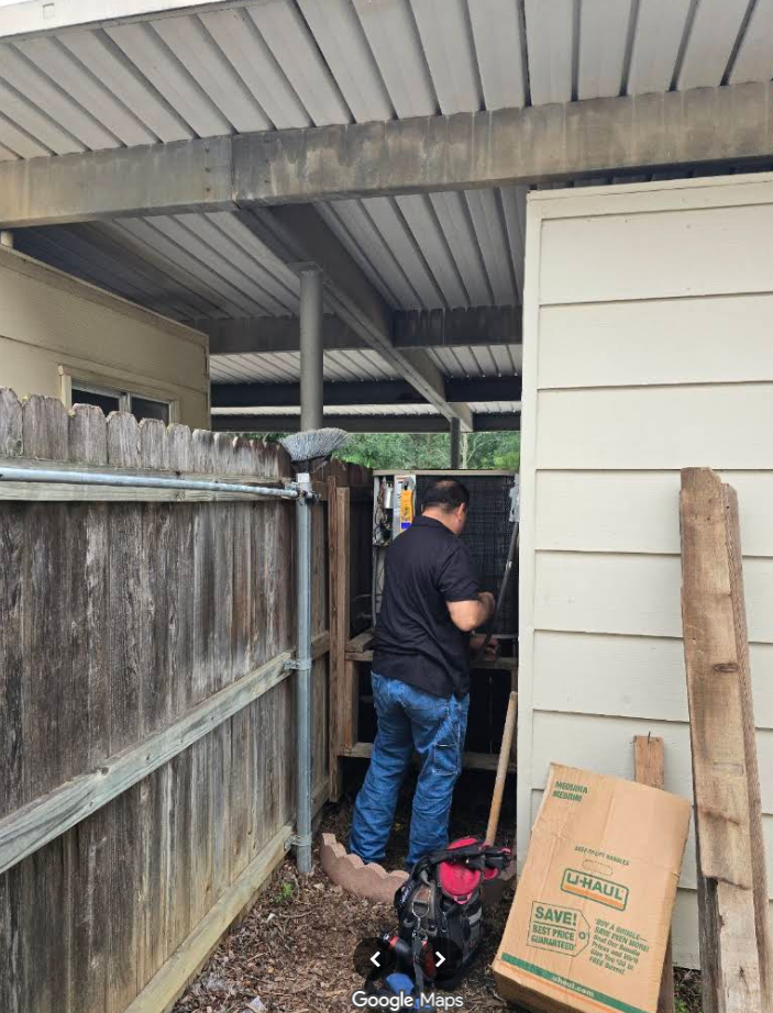 Person working in a narrow outdoor passage beside a wooden fence and white siding under a covered roof