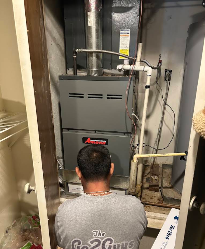 Man crouching before a gray furnace in a utility closet, with exposed pipes and wiring.