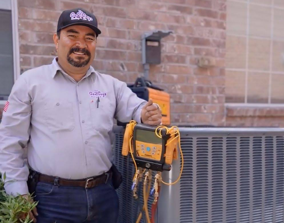 Technician holding yellow HVAC gauge set beside outdoor unit against a brick wall