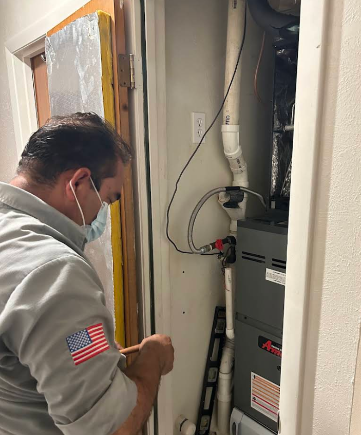 Technician inspecting HVAC unit in a narrow utility closet, wearing a mask and gray jacket.