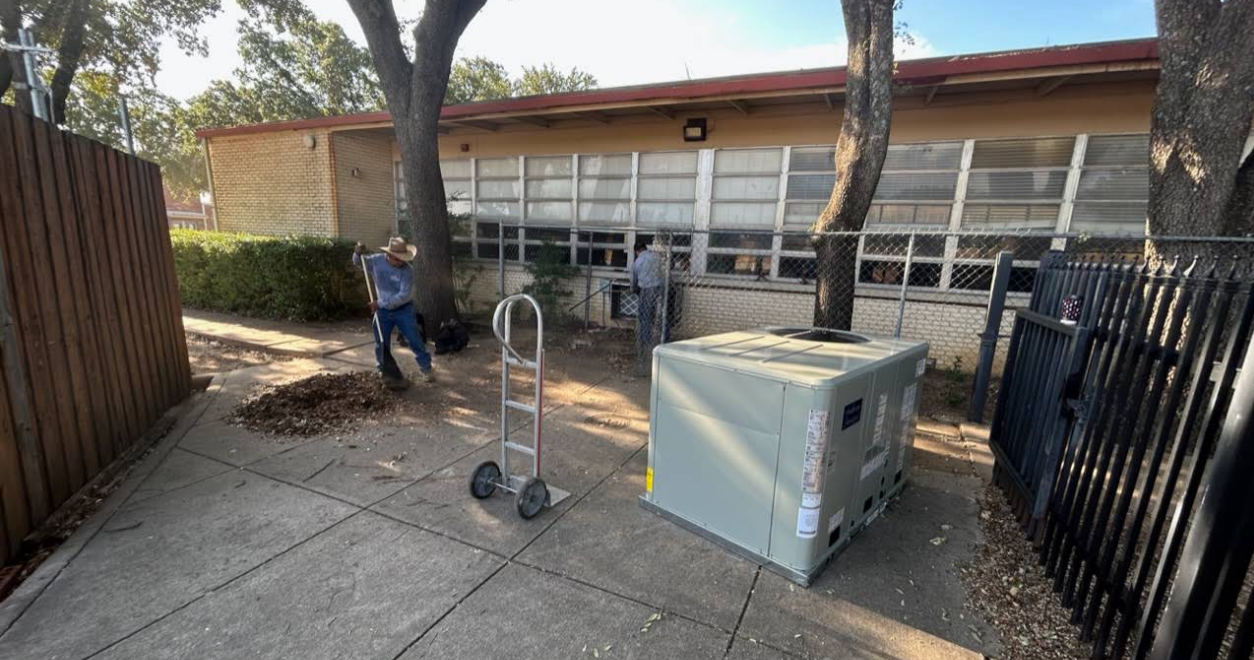 Outdoor utility area with a worker, hand truck, and large gray equipment box beside a building entrance.