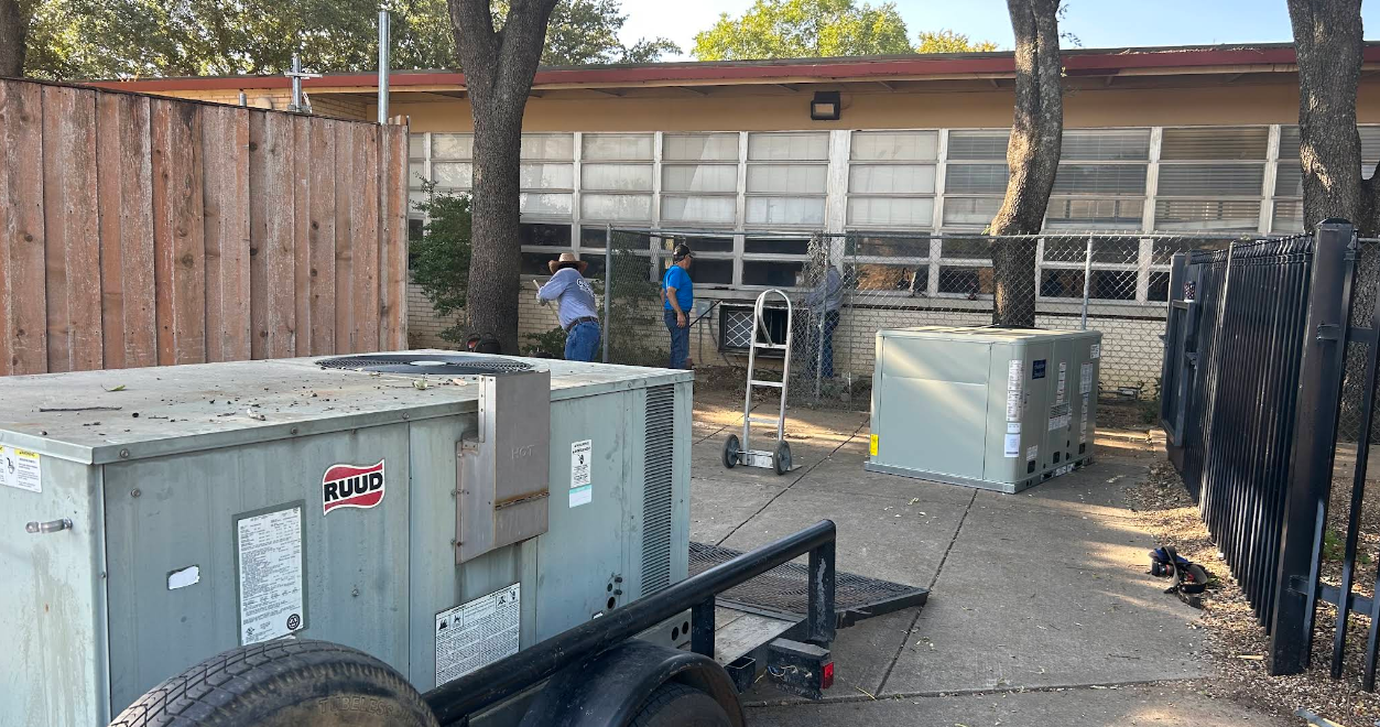 Utility boxes and trailer in a fenced yard with trees and equipment in the background