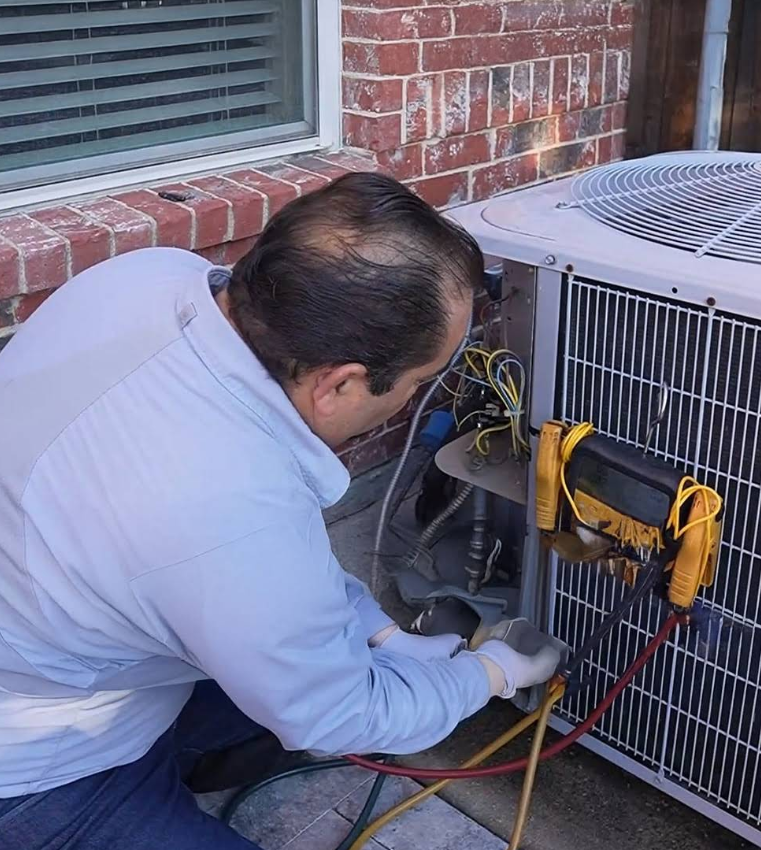 Technician servicing an outdoor HVAC unit beside a brick house, with tools and hoses attached.