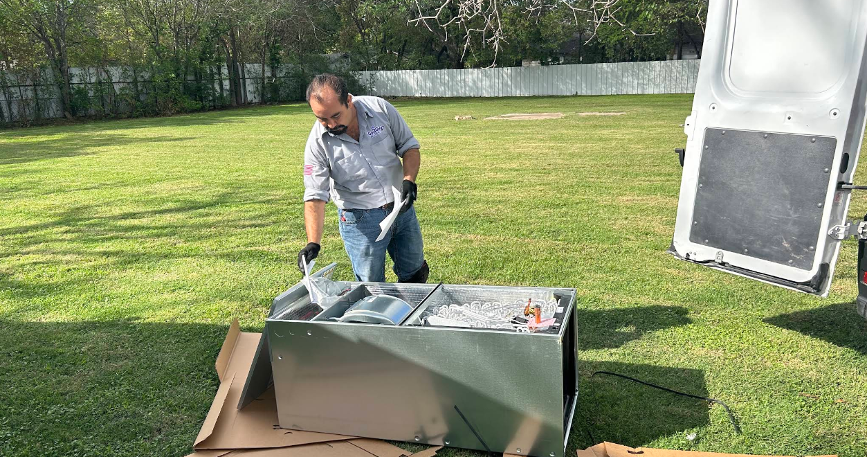 Person unloading a gray appliance from a cardboard box on a lawn outdoors
