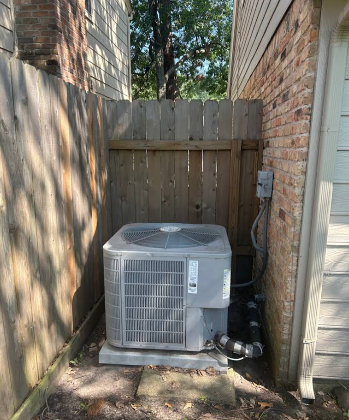 Outdoor HVAC condenser unit beside a house in a narrow fenced side yard
