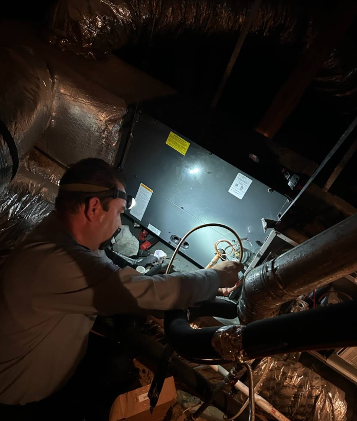 Technician inspecting HVAC equipment in a dimly lit attic, surrounded by insulated ducts.
