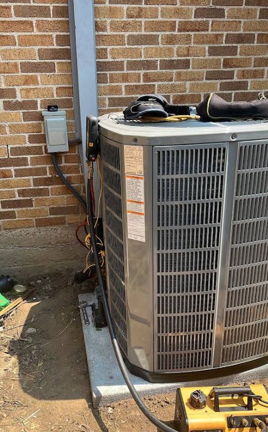 Outdoor air conditioner unit beside a brick wall with electrical conduit and a yellow tool on the ground