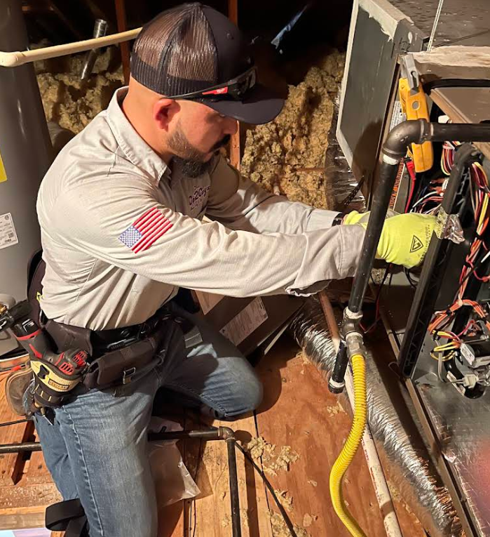 Technician kneels in a basement, adjusting plumbing lines beside an exposed wall and utility equipment.