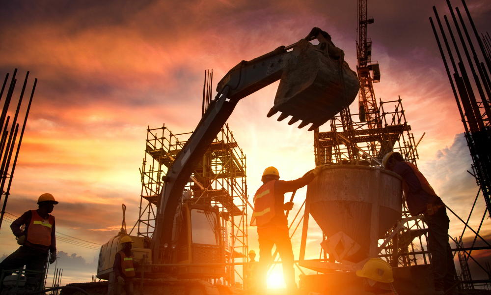 Construction workers silhouetted at sunset beside an excavator and concrete formwork on a site