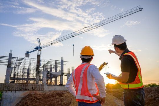 Two construction workers in safety vests and hard hats inspect a crane at a building site at sunset