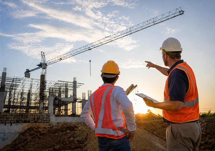 Two construction workers in safety vests and hard hats discuss a building site at sunset with a crane overhead