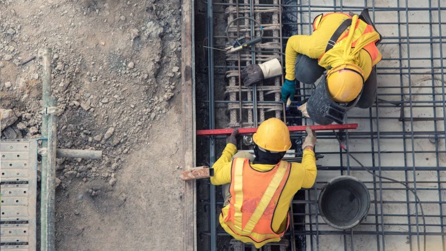 Two construction workers in yellow vests and helmets installing rebar on a concrete site.